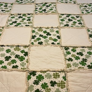 May include: A patchwork quilt with alternating squares of cream-coloured fabric and fabric printed with green shamrocks. The quilt has a frayed edge, giving it a rustic, handmade appearance. The shamrock pattern includes solid and checkered designs.