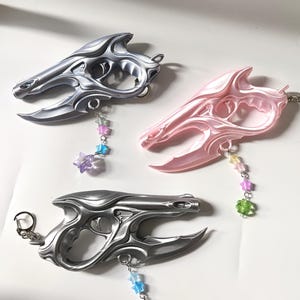 May include: Three fantasy-themed keychains in silver and pink. Each keychain features a stylized, ornate design with a loop for keys and a dangling charm of colourful beads and stars. The keychains are displayed on a white surface.