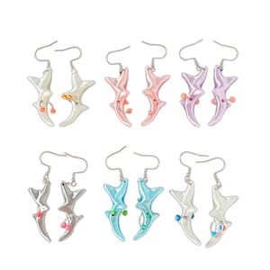 May include: A collection of six pairs of earrings in various colors. Each earring has a stylized, abstract shape with a hook and small decorative beads. Colors include white, pink, purple, silver, blue, and a pearlescent finish.