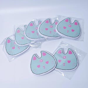 May include: A selection of cat-shaped patches. Each patch has a light blue background with pink details for the eyes, nose, and mouth. The patches are packaged in clear plastic.