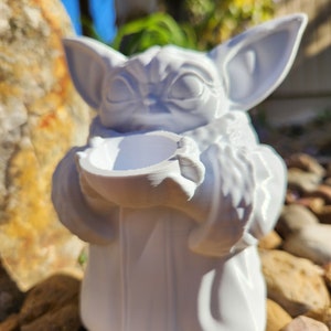 May include: A white figurine of a character with large ears and a robe, holding a small bowl. The figurine is sitting on a bed of rocks. The character's face has detailed features.