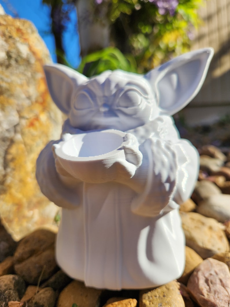May include: A white figurine of a character with large ears and a robe, holding a small bowl. The figurine is sitting on a bed of rocks. The character's face has detailed features.