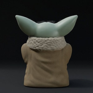 May include: A figurine of a character with large, pointed ears and a light green head. The figure is wearing a tan robe with a white, textured collar. The background is a dark gray.
