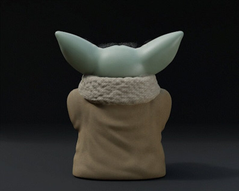 May include: A figurine of a character with large, pointed ears and a light green head. The figure is wearing a tan robe with a white, textured collar. The background is a dark gray.