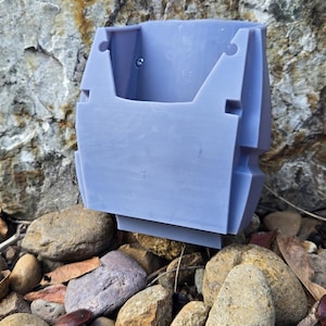 May include: A light blue, 3D-printed container with a rectangular shape and a small screw. The container is placed on a bed of rocks and pebbles, with a textured rock background. The container has a unique, geometric design.