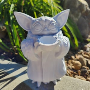 May include: A white 3D-printed figurine of a character with large ears, holding a small, round container. The figure is in a seated position, with a serene expression. The background features green foliage and a stone surface.