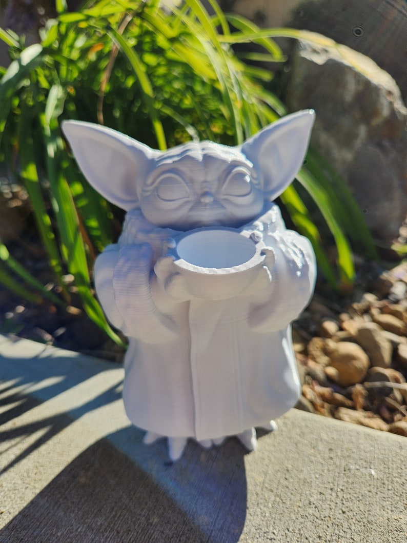 May include: A white 3D-printed figurine of a character with large ears, holding a small, round container. The figure is in a seated position, with a serene expression. The background features green foliage and a stone surface.