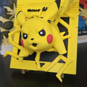 May include: A yellow Pikachu figure bursting through a yellow Nintendo 64 game cartridge. The figure has black-tipped ears, red cheek circles, and a determined expression. The text "Nintendo 64" is visible on the cartridge.
