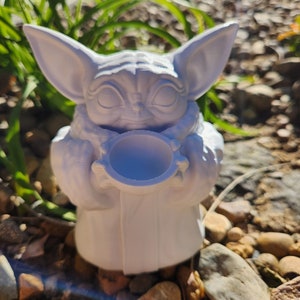 May include: A white 3D-printed figurine of a character with large ears and large eyes, holding a small bowl. The figurine is set against a background of green plants and small rocks. The character is wearing a robe.