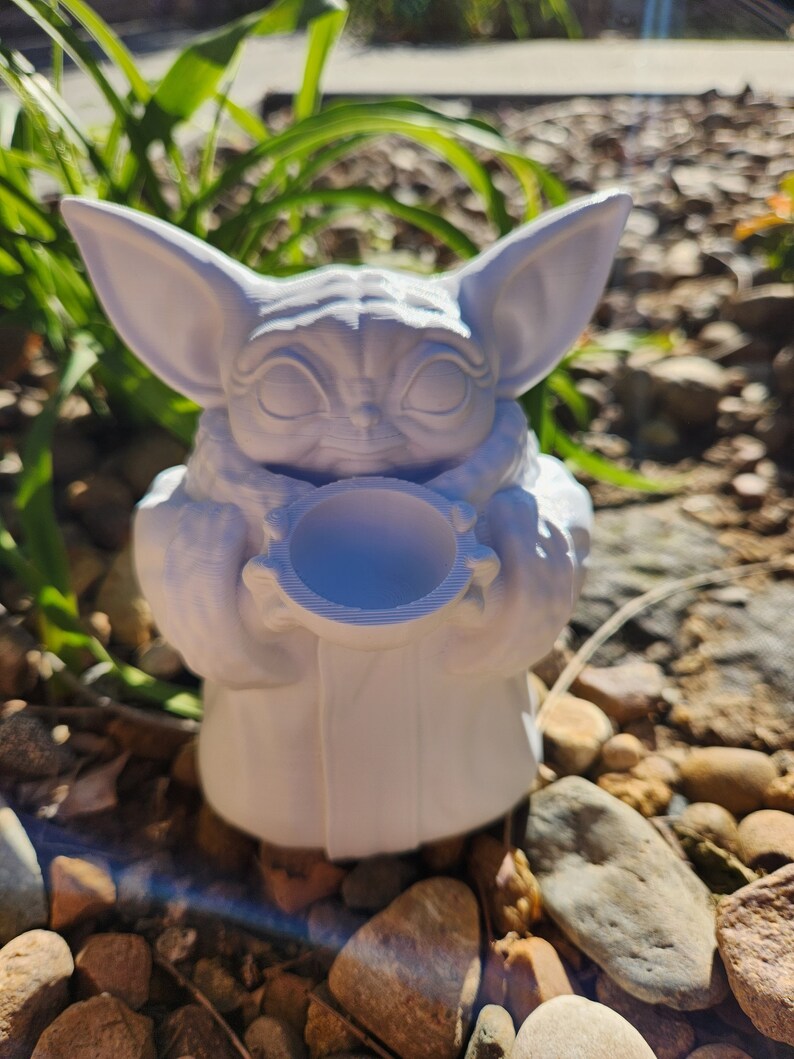 May include: A white 3D-printed figurine of a character with large ears and large eyes, holding a small bowl. The figurine is set against a background of green plants and small rocks. The character is wearing a robe.
