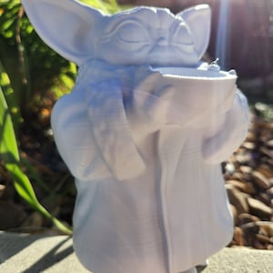 May include: A white figurine of a character with large ears and a robe, holding a bowl. The figure has closed eyes and a serene expression. The figurine is standing on a concrete surface, with greenery in the background.