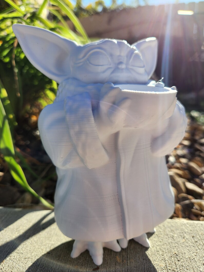May include: A white figurine of a character with large ears and a robe, holding a bowl. The figure has closed eyes and a serene expression. The figurine is standing on a concrete surface, with greenery in the background.