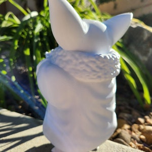 May include: A white figurine of a character with large ears and a fluffy collar. The figure is in a seated position, with a detailed texture on the collar and the folds of the garment. The figurine is set against a background of green foliage and a concrete surface.