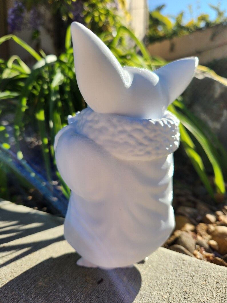 May include: A white figurine of a character with large ears and a fluffy collar. The figure is in a seated position, with a detailed texture on the collar and the folds of the garment. The figurine is set against a background of green foliage and a concrete surface.