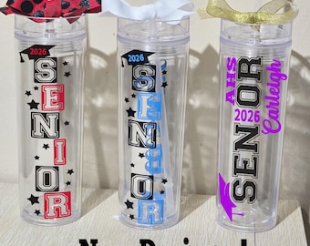 May include: Three clear plastic tumblers with straws, each adorned with a decorative bow. The tumblers are decorated with the word "SENIOR" and the year "2026", along with star accents. One tumbler has the name "Carleigh". The phrase "New Designs!" is at the bottom.