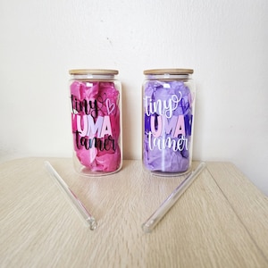 May include: Two glass jars with wooden lids and pink and purple tissue paper inside. The jars have the text "tiny uma tamer" printed on them in black with pink and purple accents. Two clear glass straws are lying on a wooden surface.