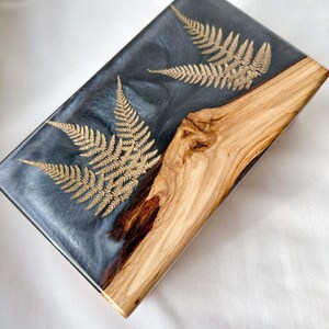 Op de afbeelding: Rechthoekig harsdienblad met een donkergrijs en zilver gemarmerd effect. Goudkleurige varenbladeren zijn ingebed in de hars. Het dienblad heeft een natuurlijk houten gedeelte aan de rechterkant, wat een rustieke touch geeft. Het is een decoratief item.