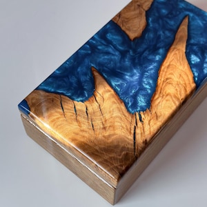 Op de afbeelding: Een rechthoekige houten doos met een glanzende afwerking. Het deksel heeft een opvallend ontwerp van natuurlijke houtnerf en een levendige blauwe harsinleg. De doos wordt waarschijnlijk gebruikt voor opslag of weergave.