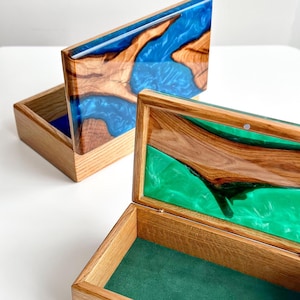 Peut inclure: Deux boîtes en bois avec des couvercles ouverts. Les couvercles présentent une combinaison de veinures de bois et de résine bleue ou verte. Les intérieurs des boîtes sont doublés de feutre bleu ou vert. Les boîtes sont en bois clair.