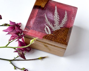 May include: A rectangular wooden box with a clear resin top. The resin is infused with a pink and purple hue and features a silver fern design. The box has a natural wood base and is decorated with a sprig of purple flowers.