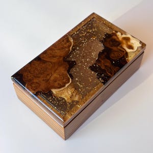 Op de afbeelding: Rechthoekige houten doos met een doorzichtige harslaag. De hars is ingebed met goudvlokken en heeft een wervelend patroon van bruin en beige hout. De doos heeft een natuurlijke houtnerf afwerking.