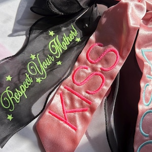 May include: A black sheer ribbon with green embroidered text reading Respect Your Mother and small star accents, paired with a pink satin ribbon featuring the embroidered word KISS in hot pink and another word in light blue thread.