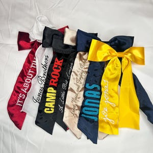 May include: Six satin hair bows with different embroidered text, including "It's About Time", "Jonas Brothers", "Camp Rock", "A Little Bit Longer", "Jonas Vines and Trying Times", "Jonas", and "Joe Jonas".