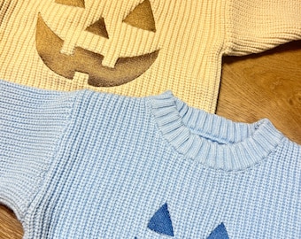 Può includere: Due maglioni lavorati a maglia, uno giallo chiaro e uno azzurro chiaro, ciascuno con un disegno a forma di faccia di zucca. I maglioni hanno polsini e colletti a coste, perfetti per Halloween. I volti delle zucche sono in colori contrastanti.