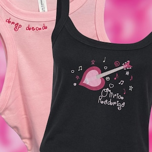 Può includere: Due canotte. Una è rosa chiaro con la scritta ricamata "dreg decade" in rosso. L'altra è nera con una grafica di chitarra a forma di cuore rosa e il nome "Olivia Rodrigo" in corsivo bianco, insieme a note musicali e stelle.