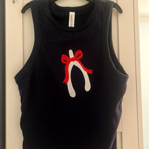 Könnte beinhalten: Ein marineblaues Tanktop mit einem weißen Glücksknochen-Design und einer roten Schleife. Das Oberteil ist ärmellos und hat einen runden Ausschnitt. Das Tanktop hängt an einem schwarzen Kleiderbügel.