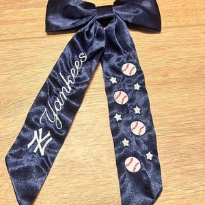 以下が含まれることがあります： ネイビーブルーのサテンヘアリボン。白い筆記体で「Yankees」の文字が入っています。リボンにはヤンキースのロゴと白い星が付いた野球ボールが描かれています。リボンは木製の表面にあります。
