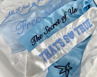 May include: A set of three blue and white satin ribbons with embroidered text. The ribbons say "All I feel is free now", "Free Now", "The Secret of Us", and "That's So True".