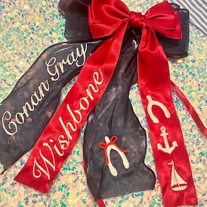 May include: A navy blue hair bow with a large red satin bow and long red ribbons. The ribbons have white embroidered text and nautical-themed designs, including a wishbone, anchor, and sailboat.