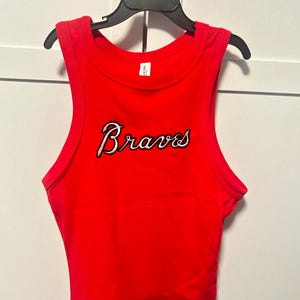 May include: A bright red tank top with the word "Braves" in white script with black outlining. The top is sleeveless and hangs on a black hanger against a white background.