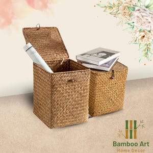 May include: Two woven storage baskets with lids, one with the lid open and a rolled up piece of paper inside. The baskets are made of natural materials and have a light brown colour. The baskets are sitting on a light brown surface. The text "Bamboo Art Home Decor" is visible in the bottom right corner of the image.