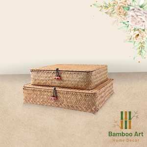 Könnte beinhalten: Zwei geflochtene, rechteckige Aufbewahrungskörbe in Naturfarbe mit Deckeln. Die Deckel haben einen schwarzen Schnurschluss und einen kleinen roten Knopf als Akzent. Die Körbe sind übereinander gestapelt. Der Hintergrund ist hellbeige.