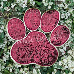 May include: A pink paw print made of sequins with a white outline. The paw print is on a green background.