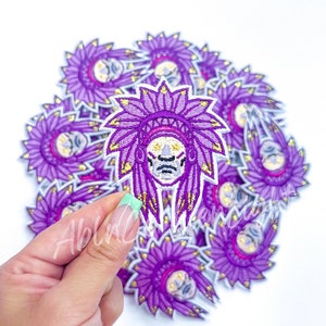 May include: A pile of purple and gold embroidered iron-on patches featuring a Native American headdress with a face inside.