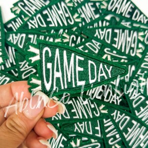May include: A pile of green felt pennant patches with white stitching that reads "GAME DAY".