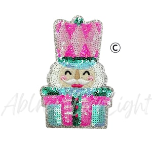 May include: A sequined patch in the shape of a nutcracker soldier. The patch features a pink and teal color scheme with a smiling face and a tall crown. The design is detailed with sequins and a white background.