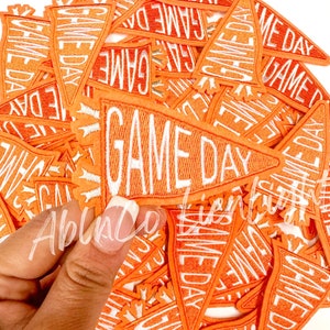May include: A pile of orange felt pennant patches with white embroidered text that reads "GAME DAY".