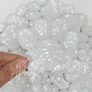 May include: A pile of white paw print patches with sequins. The patches are made of fabric and have a white outline. The sequins are white and shiny.