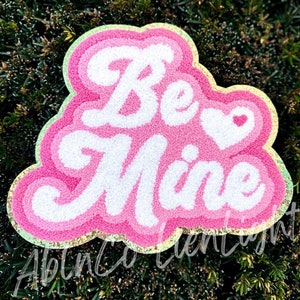 May include: A pink and white tufted rug with the words "Be Mine" in a cursive font. The rug has a heart shape with a small heart inside. The rug is surrounded by a gold and green border.