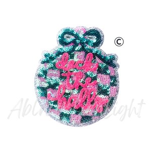 May include: A sequined ornament with a bow on top. The ornament is pink, green, and silver, with the words "deck the halls" in pink script. The background is white.