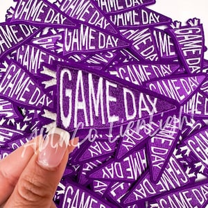 May include: A collection of purple pennant-shaped patches with the words "GAME DAY" embroidered in white. The patches have a white fringe along the edges. One patch is being held by a hand.