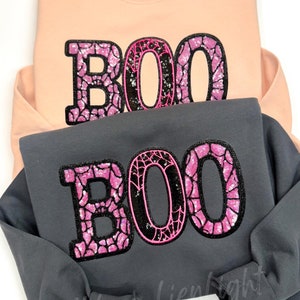 May include: Two sweatshirts with the word "BOO" in pink and black sparkly letters. The letters are outlined in pink and have a spiderweb design.