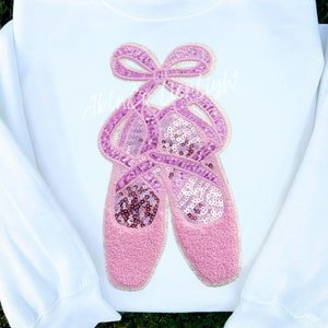 May include: White sweatshirt with a pink sequined ballerina shoe design. The shoes are tied together with a pink bow.