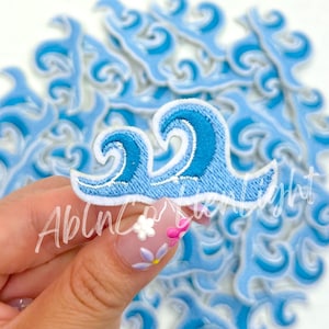 May include: Embroidered wave patches in shades of blue and white. The patches feature a detailed design of ocean waves, with a light blue base and darker blue accents. The patches are ideal for adding a nautical touch to clothing or accessories.