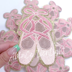 May include: Pink and gold embroidered ballet shoe patches. The shoes are tied together with a pink ribbon and bow.