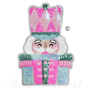 May include: A sequined Christmas ornament in the shape of a nutcracker soldier. The ornament features a pink and blue color scheme with silver accents. The nutcracker has a smiling face and is wearing a tall hat.